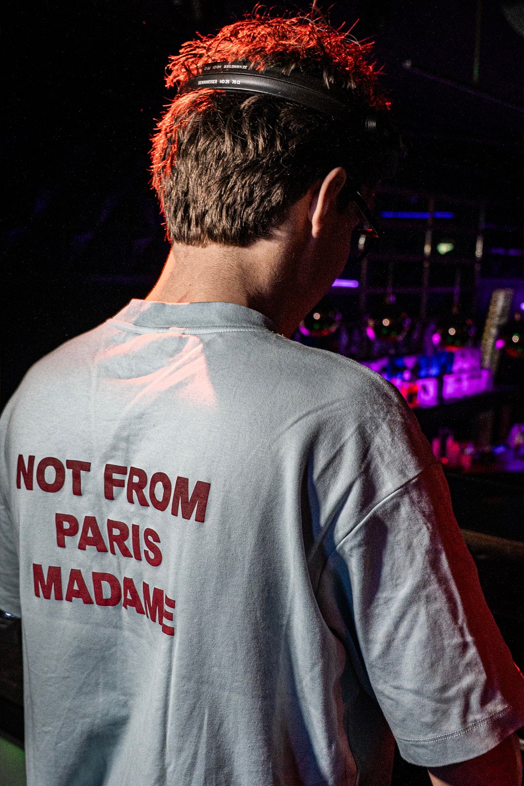 FØR U DJ seen from behind in a "Not From Paris Madame" T-shirt, headphones on, club lights ahead