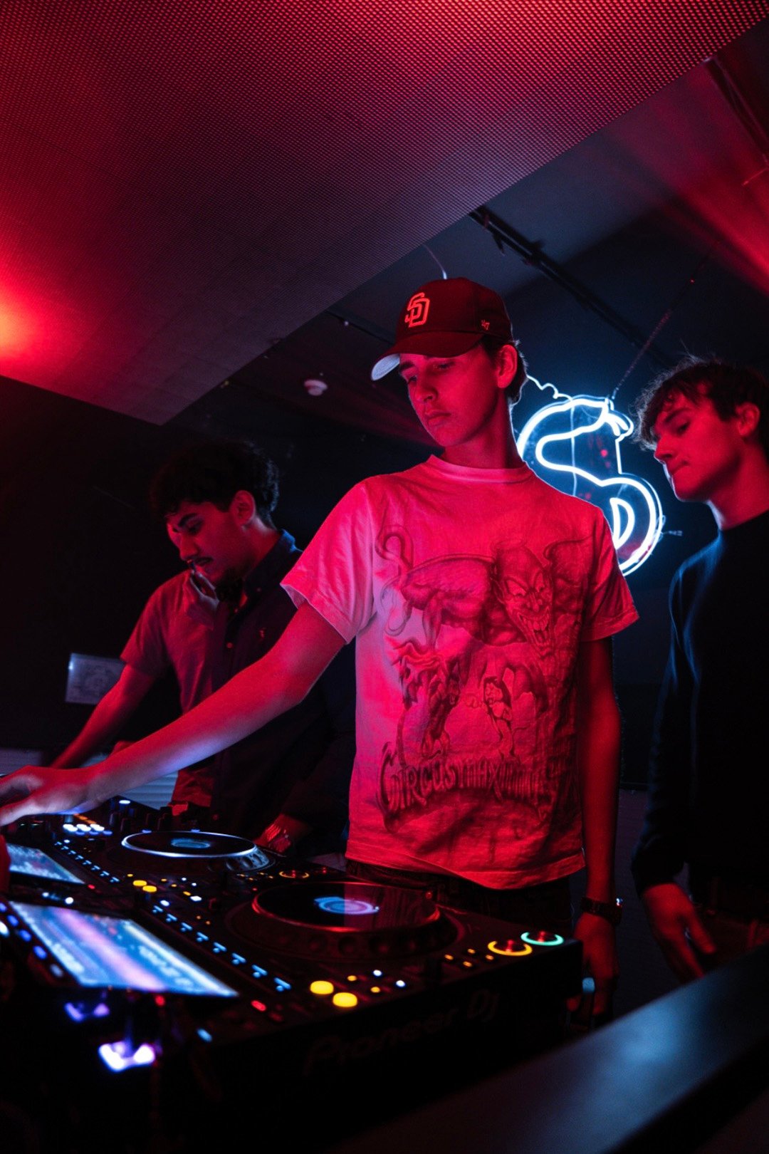 FØR U DJ in a cap mixing on the decks under red light, a neon sign and friends behind him