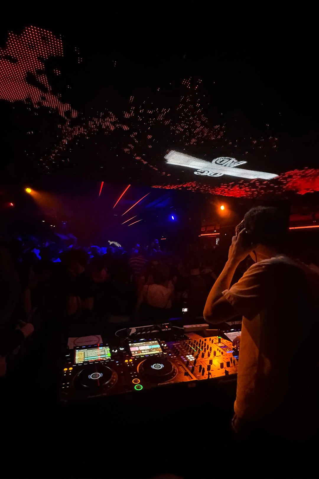 FØR U DJ in headphones looking out from the booth over a dark club dance floor and light show